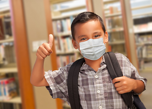 Hispanic Student Boy Wearing Face Mask with Thumbs Up and Backpack in the Library.