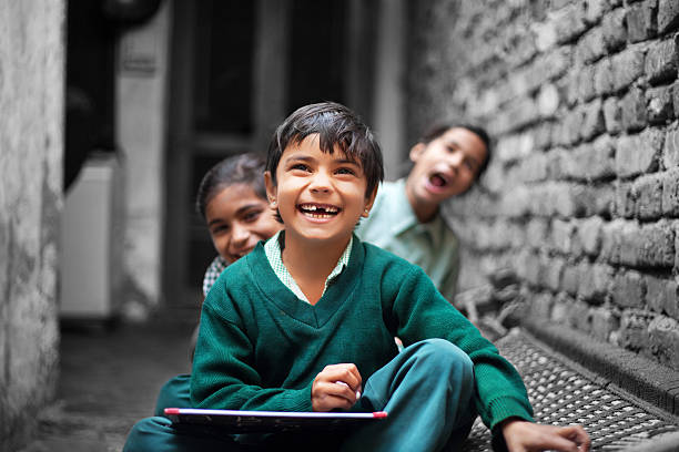 Cheerful School Girls of Indian Ethnicity sitting in a row on the Charpai near brick wall at home wearing school dress & Laughing together. One of them Holding slate(Chalkboard) & writing with the chalk, they all are laughing together and looking to the camera while sitting on Charpai (Domestic Bed) at home in Rural India.