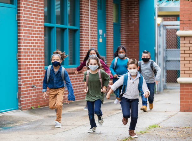 A group of six multi-ethnic elementary school students running outside the school building, carrying backpacks, and wearing face masks. They are back to school during the covid-19 pandemic. The boys and girls are 7 to 10 years old.